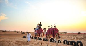 An Arab man at Dubai Desert Safari holding a camel with some people on it.